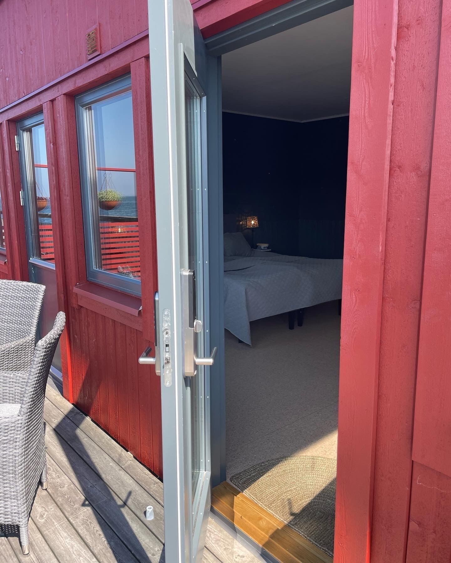 A room with a bed, a nightstand, and a window overlooking a balcony with wicker furniture.