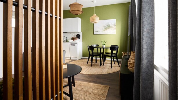 Une cuisine moderne avec des meubles blancs, un coin repas avec des chaises noires et un mur vert orné d’un tableau encadré.