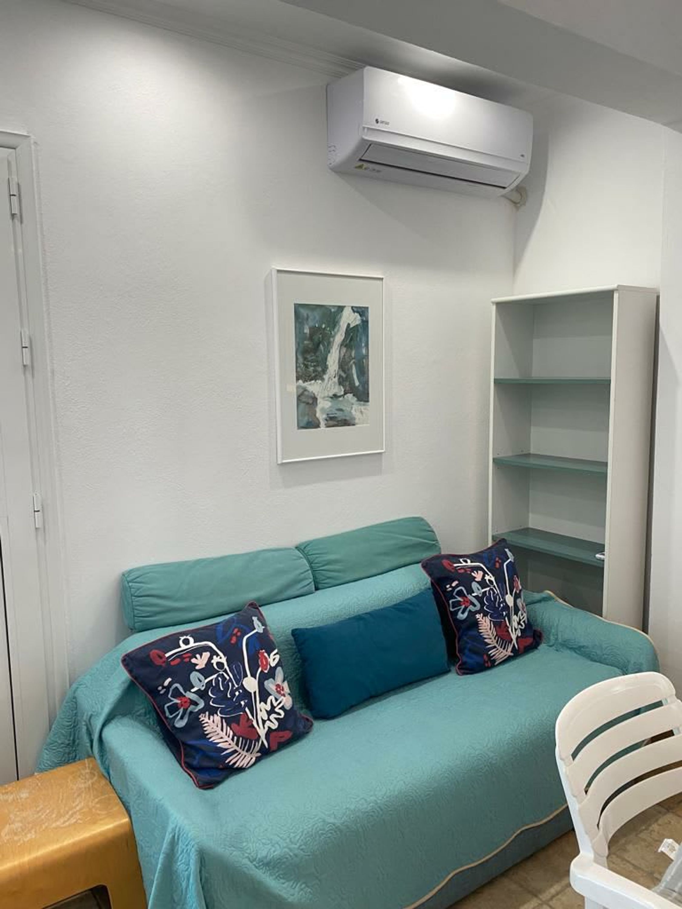 A room with a teal sofa, two patterned pillows, a framed picture, a white air conditioning unit, and a white shelving unit.