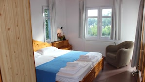 Een slaapkamer met een houten bed, een blauwe spreidek, een stoel, een nachtkastje met een lamp en een raam met uitzicht op groen.