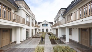 A courtyard with a central pathway flanked by two-story buildings with balconies, wooden doors, and a central garden area. - Dünenloft, House Kliff Whg. 11 (Sylt)