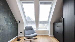 Een kamer met een schuine balkenlaag, twee dakramen, een grijze stoel, een zwarte vloerlamp en een donkerhouten dressoir.