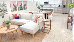 Un salon moderne comprenant un canapé d’angle, un coin repas et une cuisine avec des meubles blancs et des appareils électroménagers en acier inoxydable.