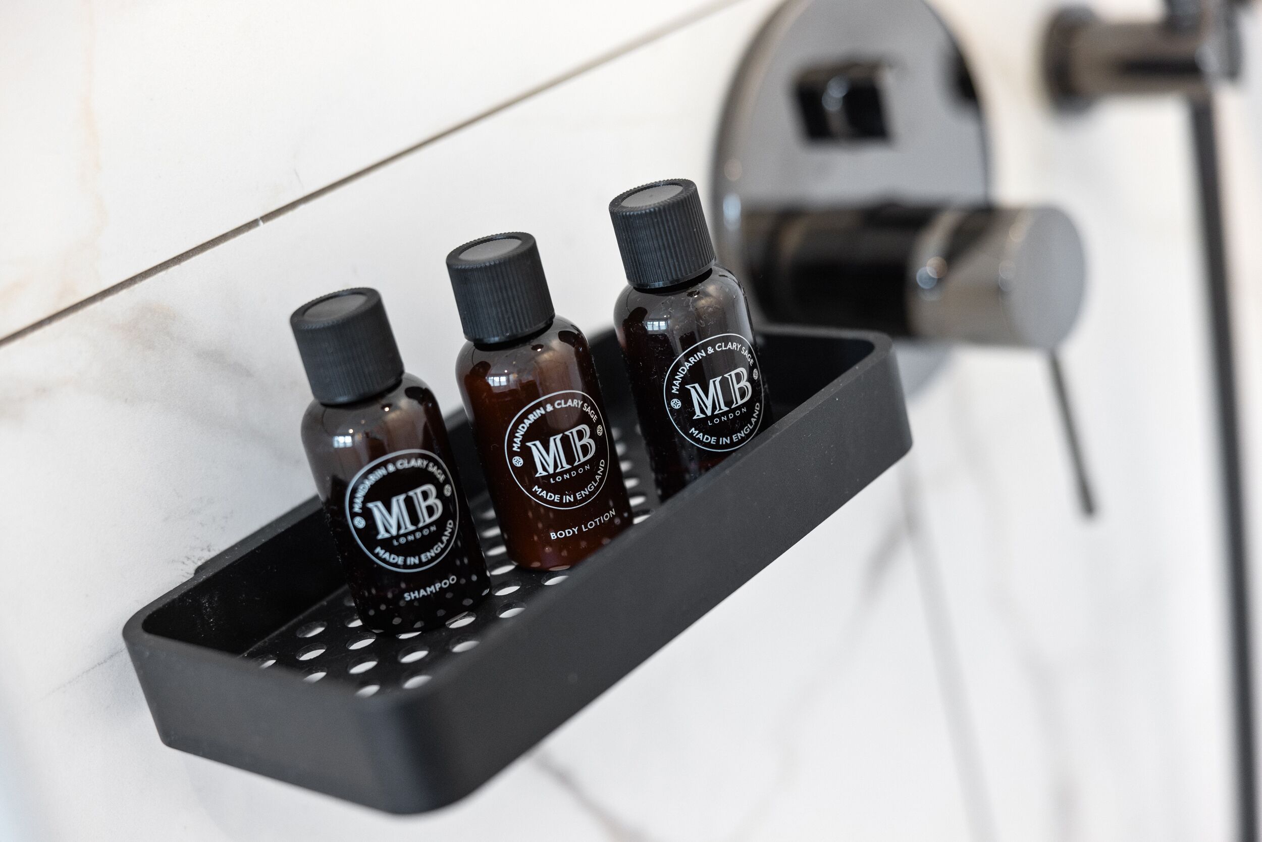 A black tray holding three bottles of MUB body lotion and shampoo, with a bathroom faucet in the background.