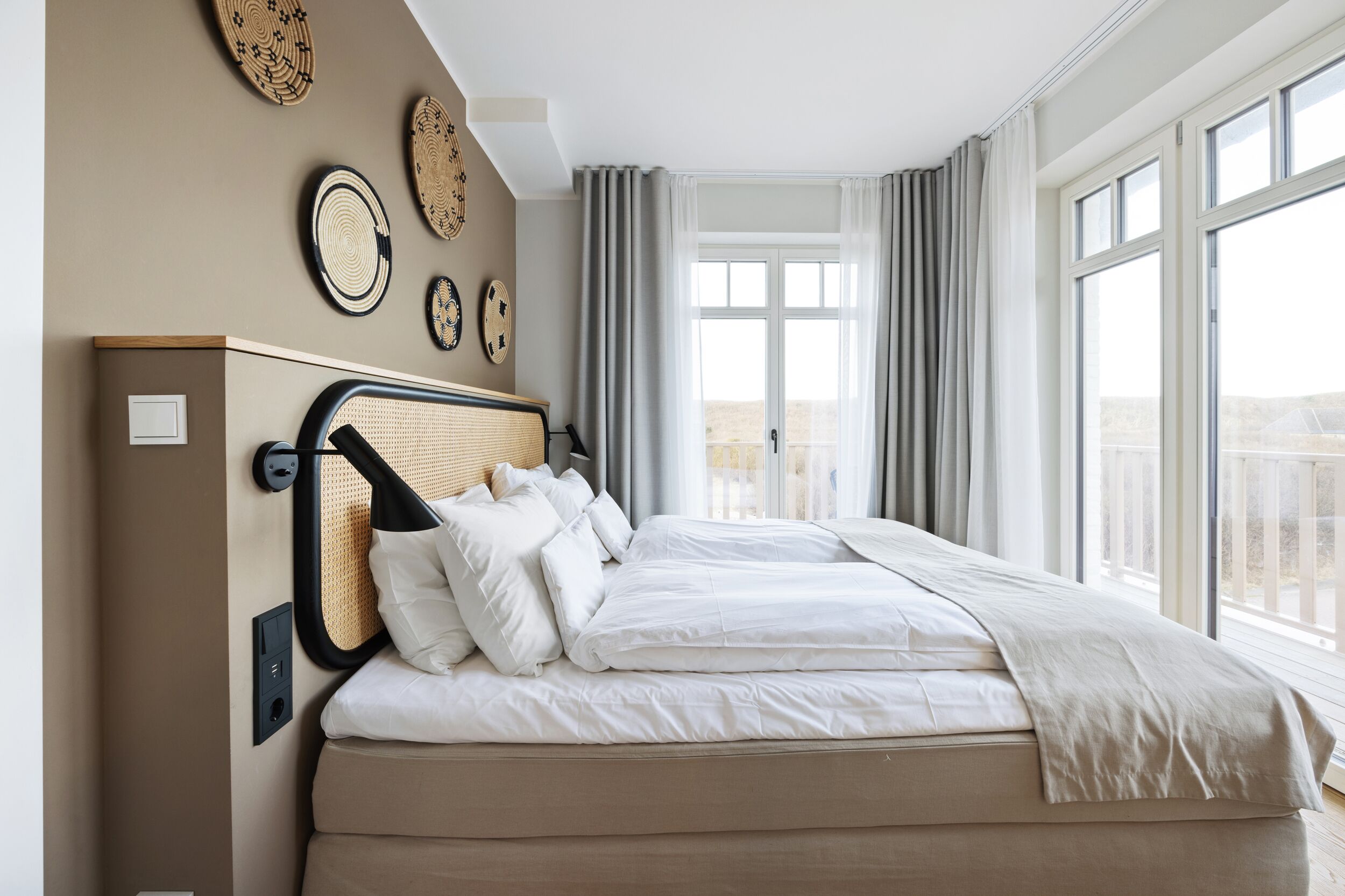 A neatly made bed with white linens and a beige headboard, a large window with curtains, and wall decor including woven plates.