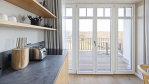 A modern kitchen with a built-in toaster, wooden shelves, and a large sliding glass door leading to an outdoor deck. - Dünenloft, House Kliff, Whg. 9 (Sylt)