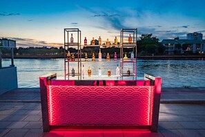 A waterfront bar with multiple shelves of bottles, a red illuminated seating area, and a calm river in the background. - Sunny Oasis | Swimming. Restaurant (Hollywood)