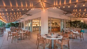 An outdoor dining area with round tables and chairs, ceiling fans, and string lights. - Sunny Retreat | Restaurant + Outdoor Pool (Hollywood)