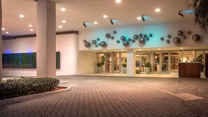 A modern hotel entrance with a glass facade, a reception desk, and a landscaped area with a brick pathway. - Sunny Retreat | Restaurant + Outdoor Pool (Hollywood)