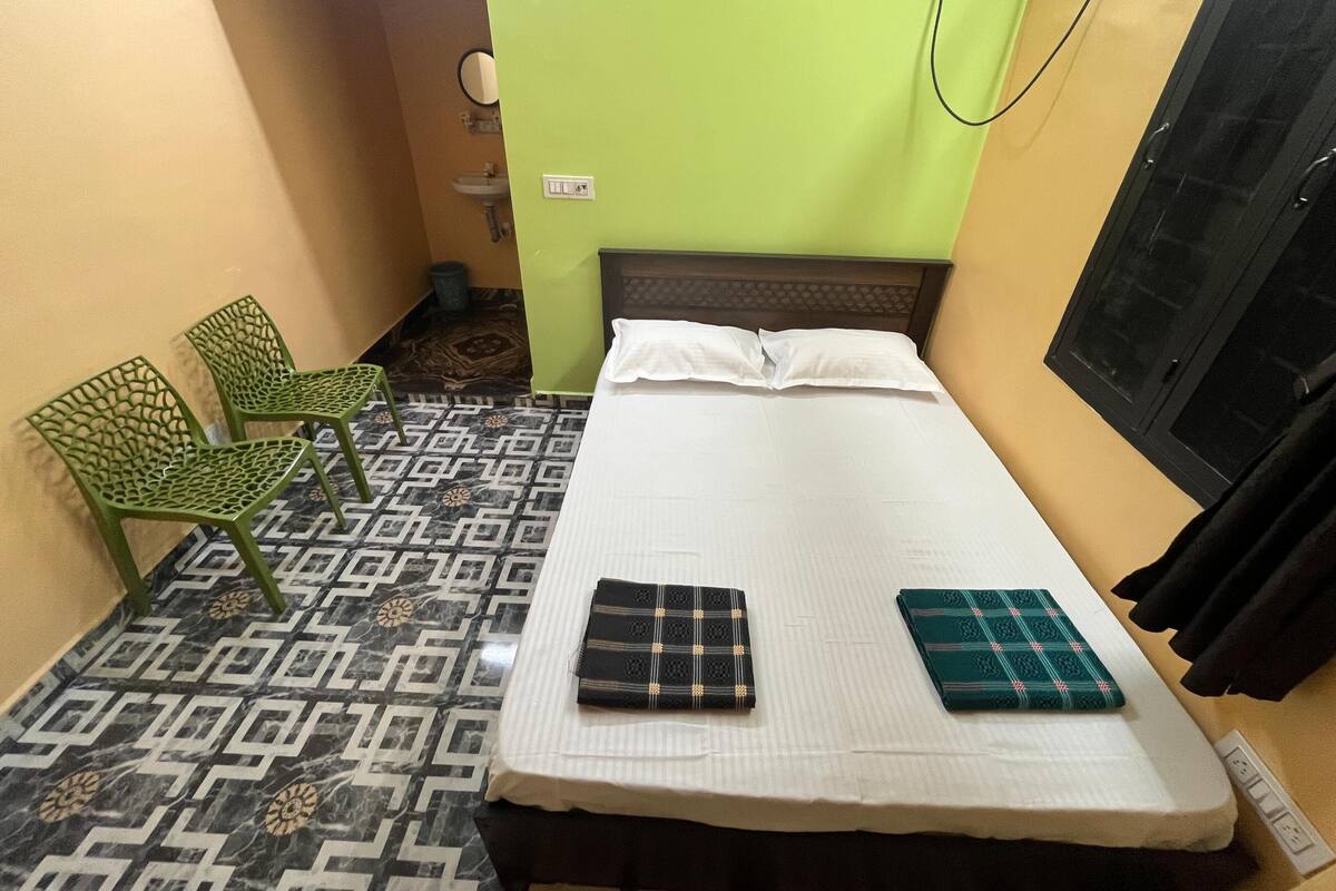 A small, single-bed room with a patterned floor, a green chair, and a bedside table with two folded blankets.