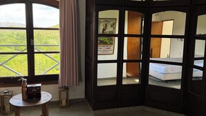 A room with a bed, a table with a vase and books, a window with a view of greenery, and a door with a poster. - Bell Vivere Guesthouse (El Volcan)