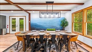 Ein Essbereich mit großem Tisch, gedeckt für sechs Personen, Holzbalken an der Decke und Bergblick durch ein Fenster.