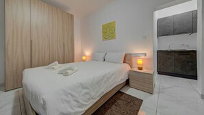 A modern bedroom with a wooden wardrobe, a bed with white linens, a small bedside table with a lamp, and a kitchenette area with a sink and cabinets. - Bright Msida Studio · King Bed - Fast WiFi (Msida)