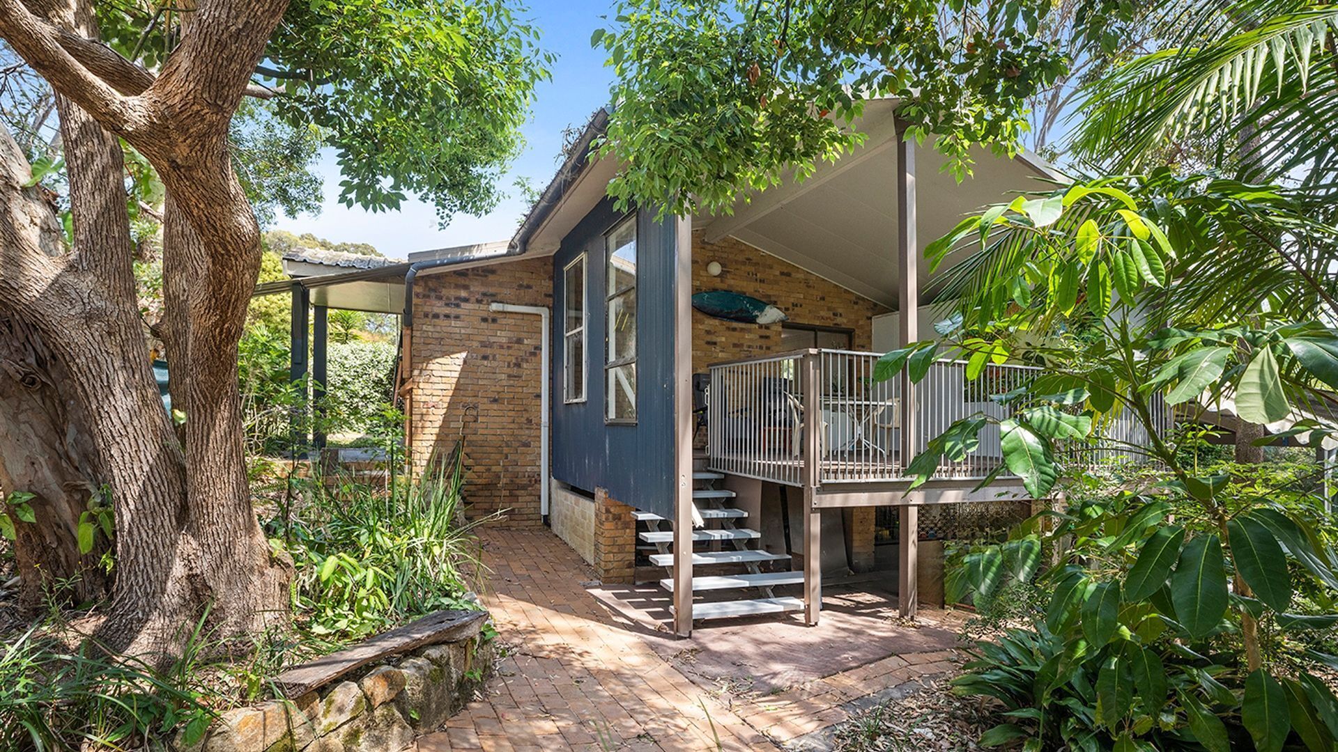 Une maison avec un escalier menant à un balcon, entourée de verdure et d’un arbre.