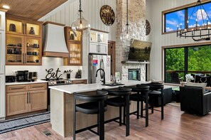 A modern kitchen with a central island, bar stools, a stone fireplace, and large windows. - Beneath the Stars | Luxury 3BR Cabin w/ Indoor Pool & Game Room (Gatlinburg)