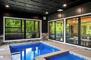 A swimming pool with stone walls and large windows, a ceiling with recessed lighting, and a view of greenery outside. - Beneath the Stars | Luxury 3BR Cabin w/ Indoor Pool & Game Room (Gatlinburg)