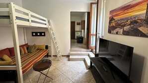 Een knus woongedeelte met een stapelbed, een slaapbank, een televisie en een schilderij van een stadsgezicht.