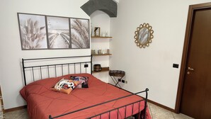 Een slaapkamer met een metalen bedframe, een rode sprei en twee kussens. Er zijn wandplanken met decoratieve items en een spiegel.