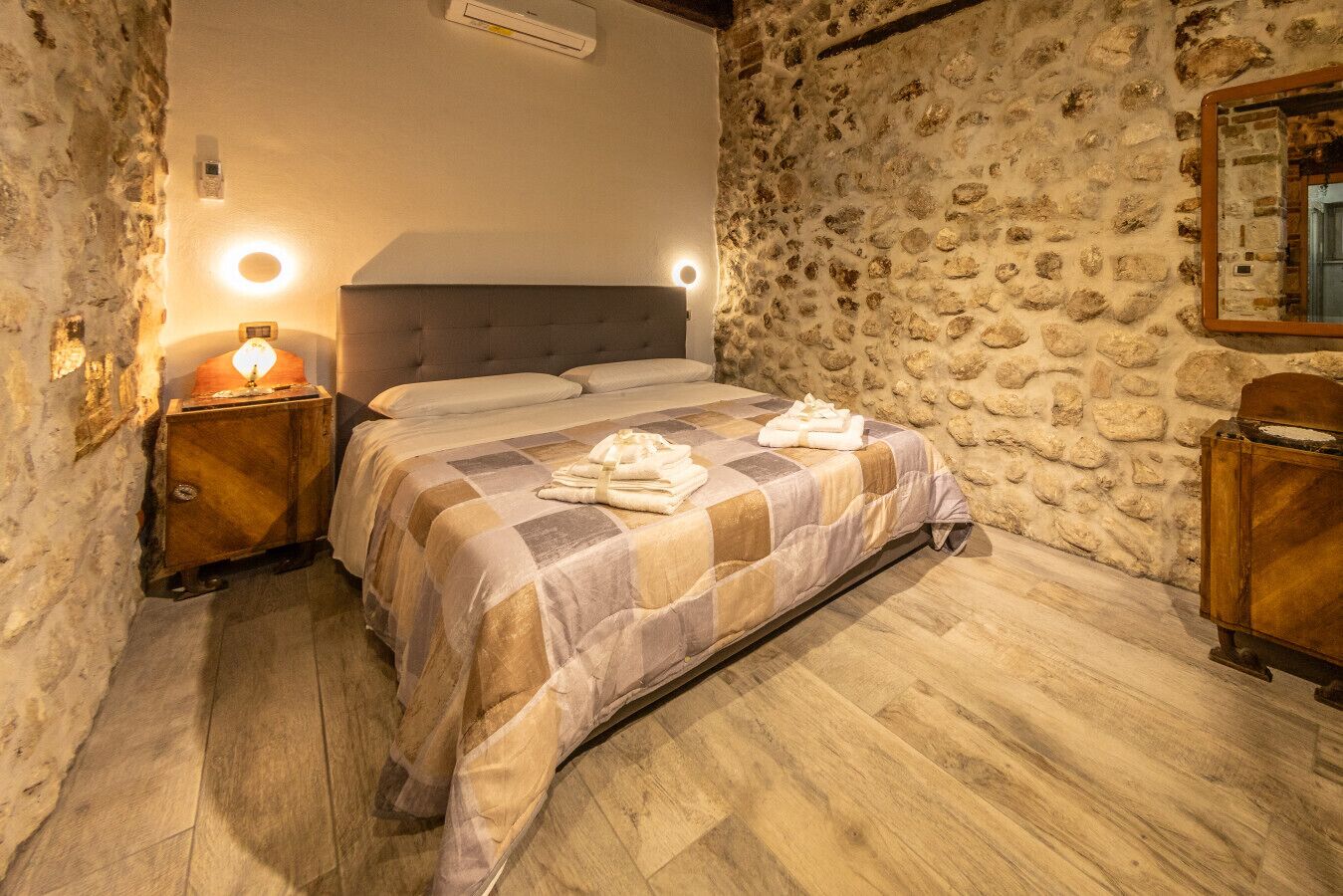Een slaapkamer met een stenen muur, een houten bed, een nachtkastje met een lamp en een spiegel aan de muur.