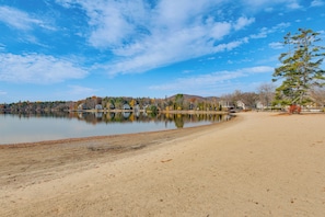 A serene lakeside scene with a sandy beach, calm water, and a row of houses in the background. - < 1 Mi to Lake Winnipesaukee: Family Winter Escape (Moultonborough)