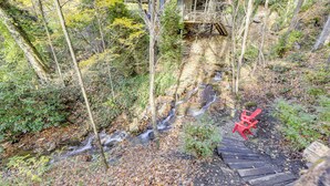 A wooden staircase leading down to a stream in a forested area with a red chair on the path. - Creekside Cabin in the Heart of Maggie Valley (Maggie Valley)
