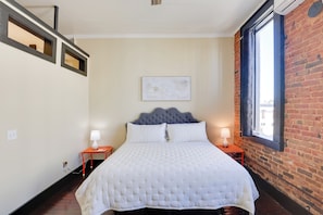 A bedroom with a large bed, two bedside tables with lamps, a window with a view, and a brick wall. - Convenient Couple's Retreat in Dtwn Marshall! (Marshall)
