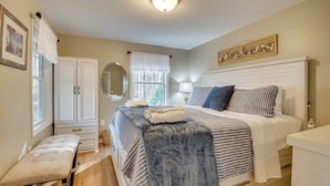 Une chambre à coucher comprenant un lit, des tables de chevet, un banc, une armoire, un miroir et une fenêtre avec des rideaux.