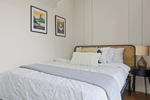 Une chambre à coucher avec un lit, deux tableaux encadrés au mur, une lampe de chevet et une tête de lit en osier.