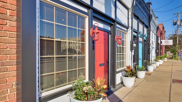 A row of buildings with large windows and doors, some with shutters, and potted plants along the sidewalk.