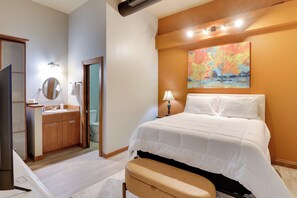 A hotel room with a bed, a bedside table with a lamp, a bathroom with a sink and mirror, and a painting on the wall. - Ultimate Convenience: Historic Dtwn Marshall Apt! (Marshall)