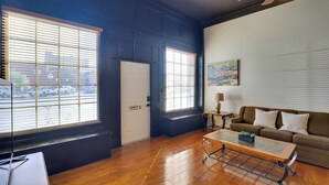 A spacious living room with a wooden floor, a brown sofa, a coffee table, and large windows with blinds. - Steps to Restaurants & Shops! Dtwn Marshall Apt (Marshall)