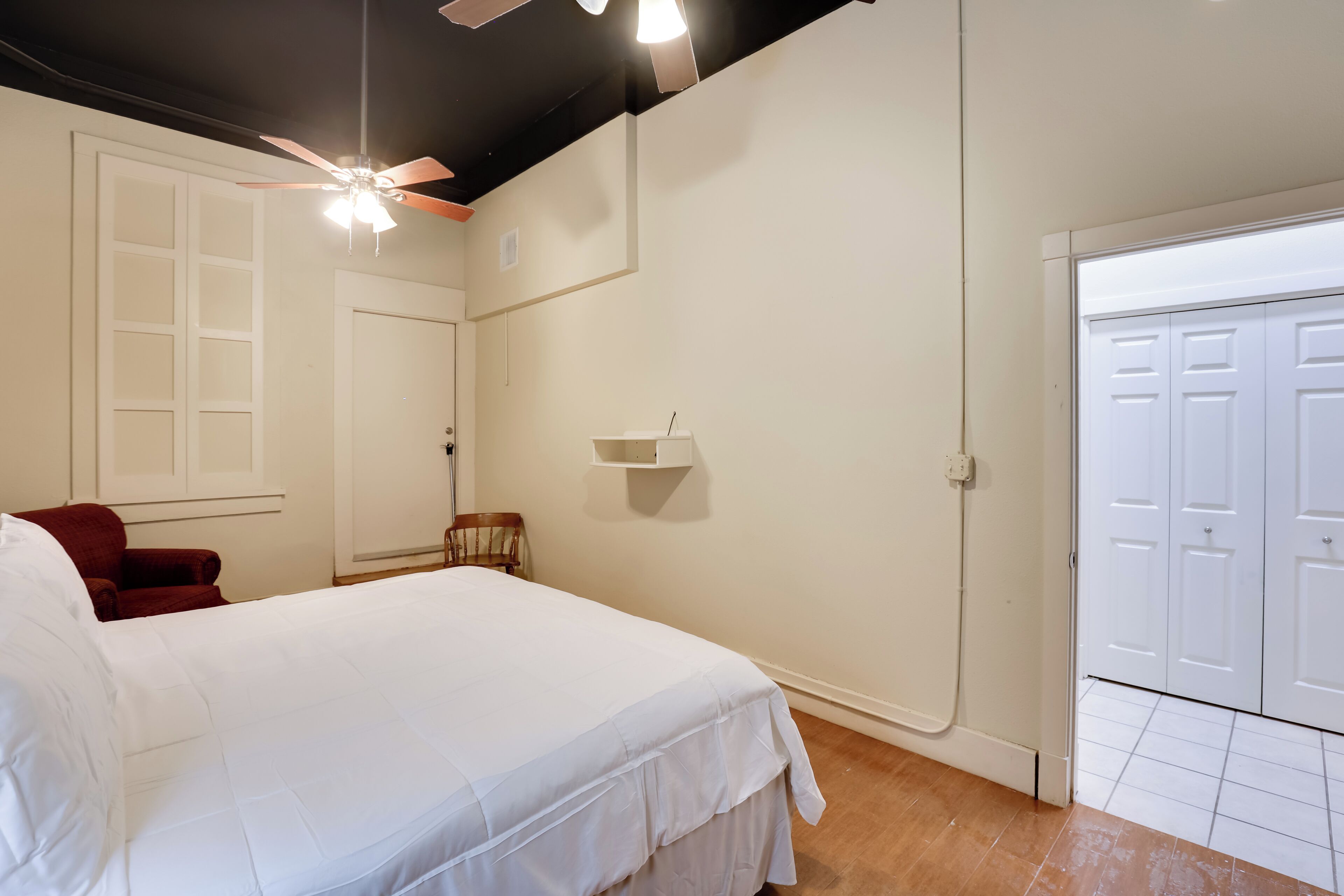 Un dormitorio con una cama grande, un ventilador de techo, una silla roja y un armario blanco.