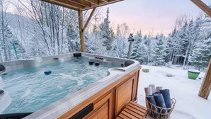 Un jacuzzi en un entorno nevado al aire libre con una terraza de madera y una cesta de toallas.