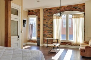Una habitación con pared de ladrillo, una cama, una mesa, una silla y una ventana con cortinas.