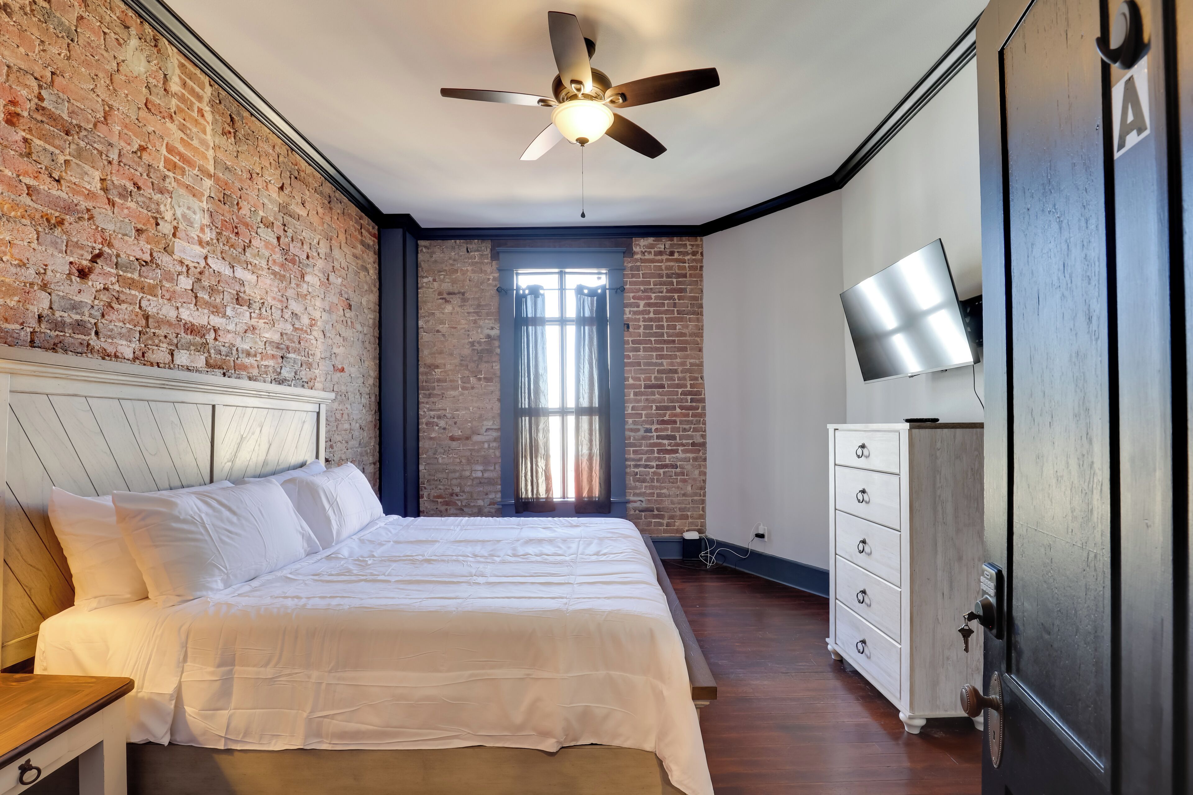 Un dormitorio con pared de ladrillo, una cama con ropa de cama blanca, un ventilador de techo, un aparador y un televisor en la pared.