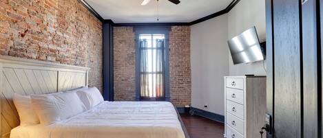 Un dormitorio con pared de ladrillo, una cama con ropa de cama blanca, un ventilador de techo, un aparador y un televisor en la pared.