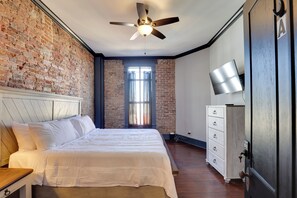 A bedroom with a brick wall, a bed with white bedding, a ceiling fan, a dresser, and a TV on the wall. - Walk to Museums! Historic Downtown Marshall Apt (Marshall)