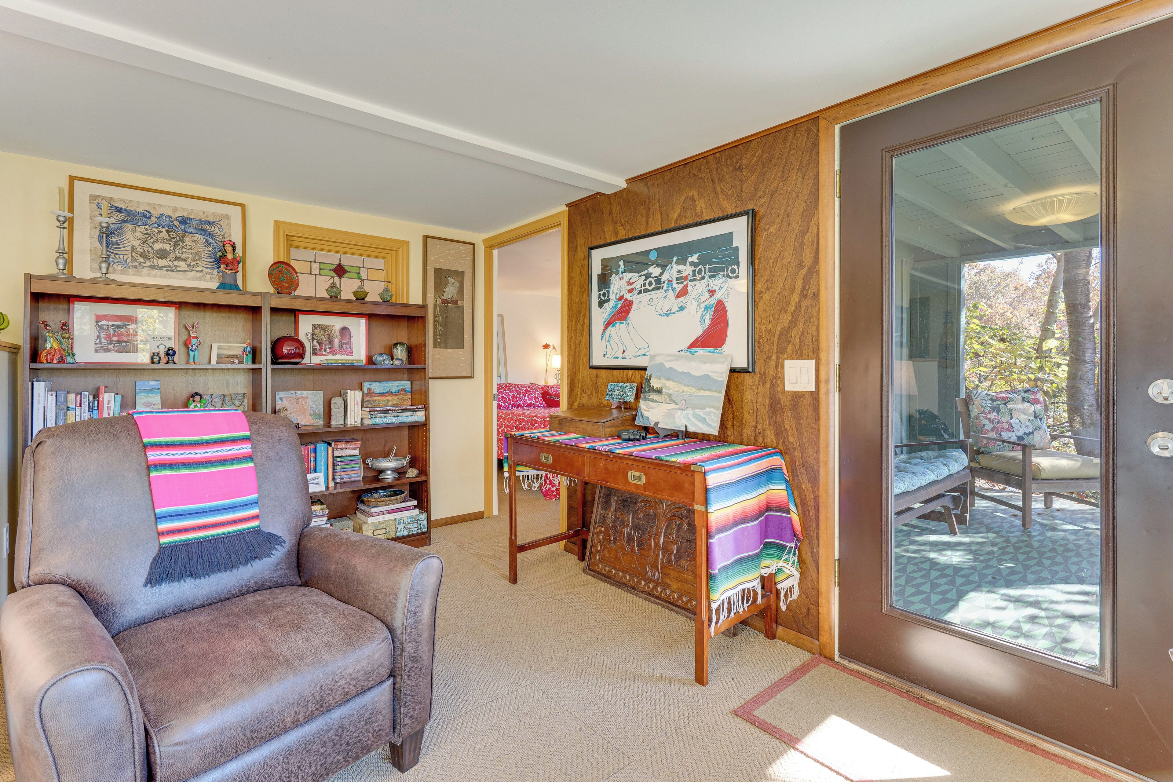 Una sala de estar acogedora con un sillón de cuero, un escritorio de madera con una manta colorida, una estantería llena de libros y adornos, y un ventanal con vistas al exterior.