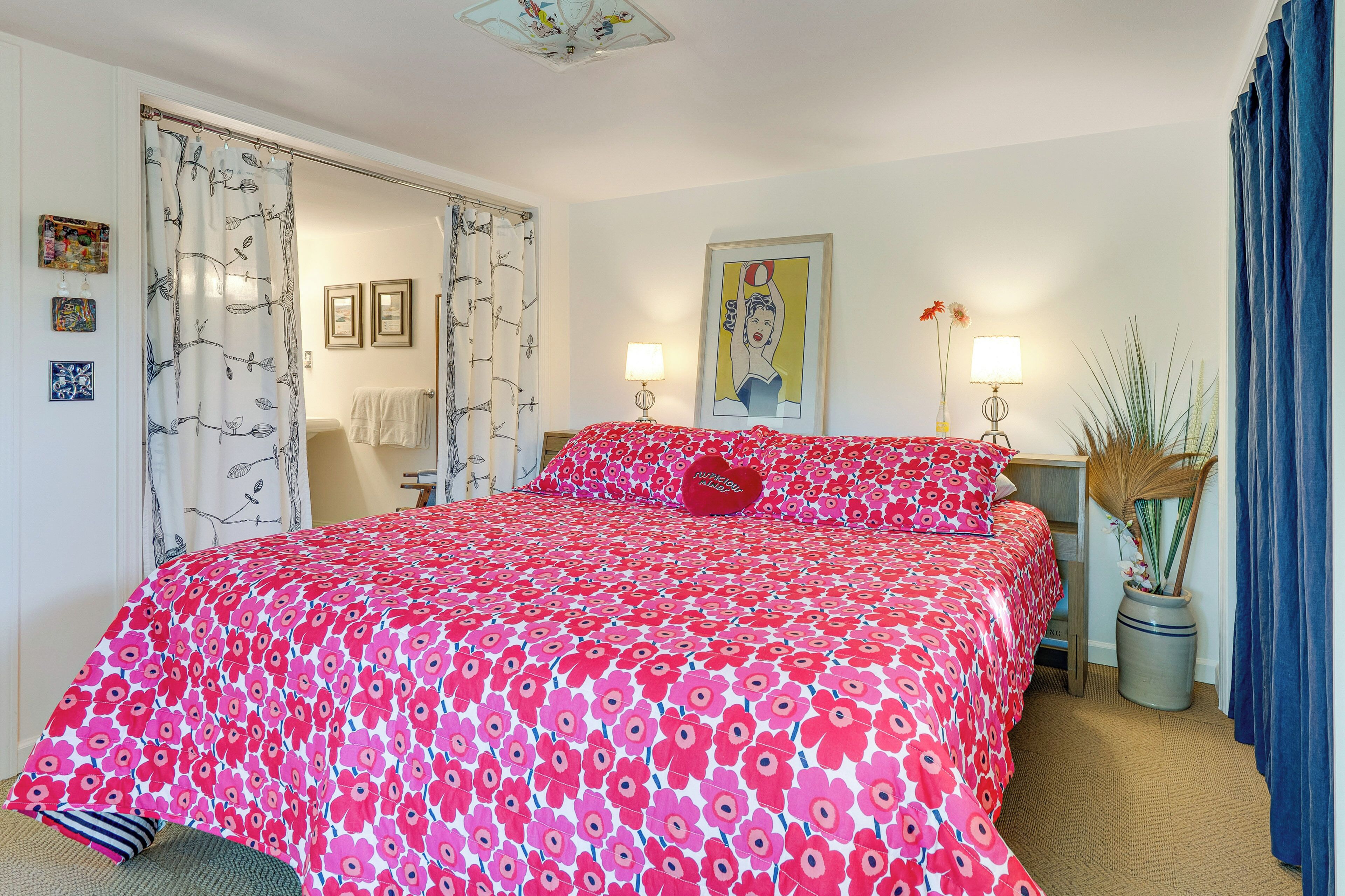 A bedroom with a large bed, two bedside tables with lamps, a mirror, a potted plant, and a shower curtain with a leaf pattern.