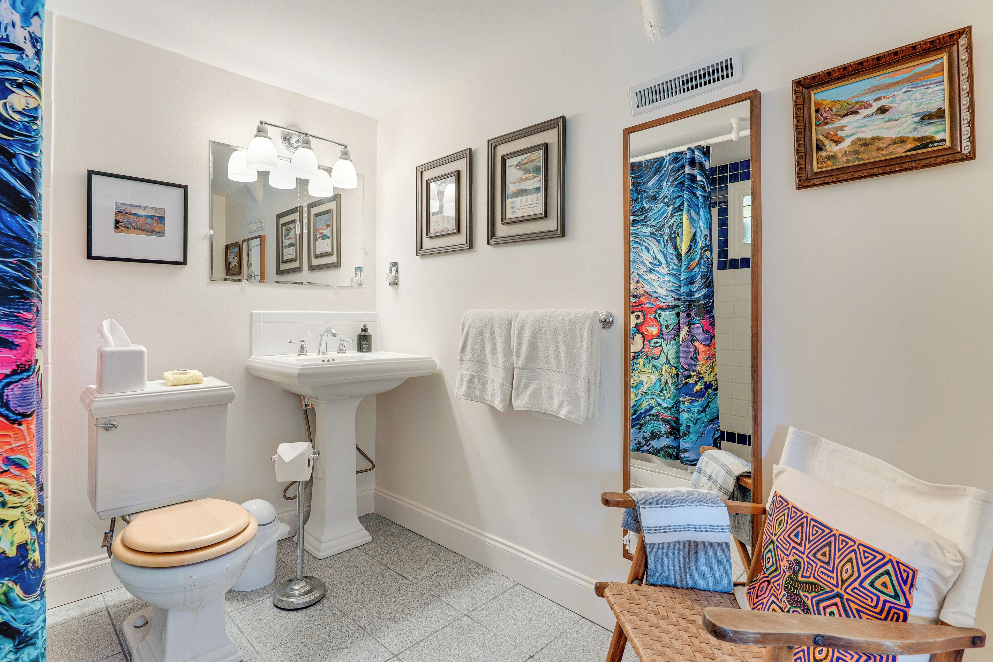 A bathroom with a toilet, sink, and mirror. There are towels and a chair with a colorful cushion.
