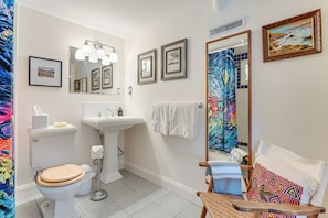 Baño con inodoro, lavabo y espejo. Hay toallas y una silla con un cojín colorido.
