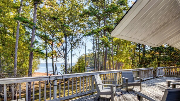 A wooden deck with a railing, two chairs, and a table overlooking a beach and trees. - Dock + Views: Family Retreat on Lake Hartwell! (Townville)