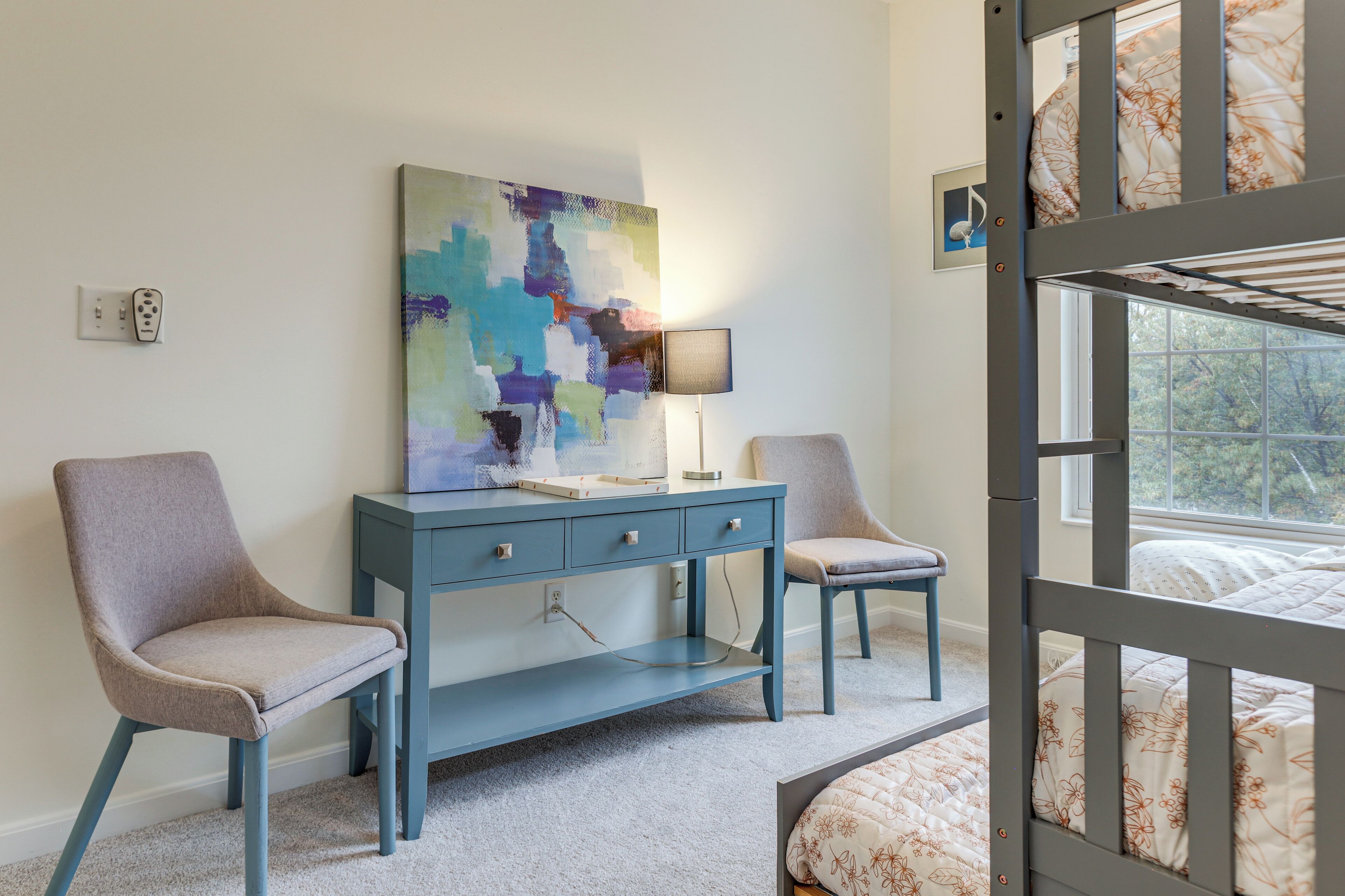 A bedroom with a bunk bed, a desk with chairs, a colorful abstract painting, and a lamp.
