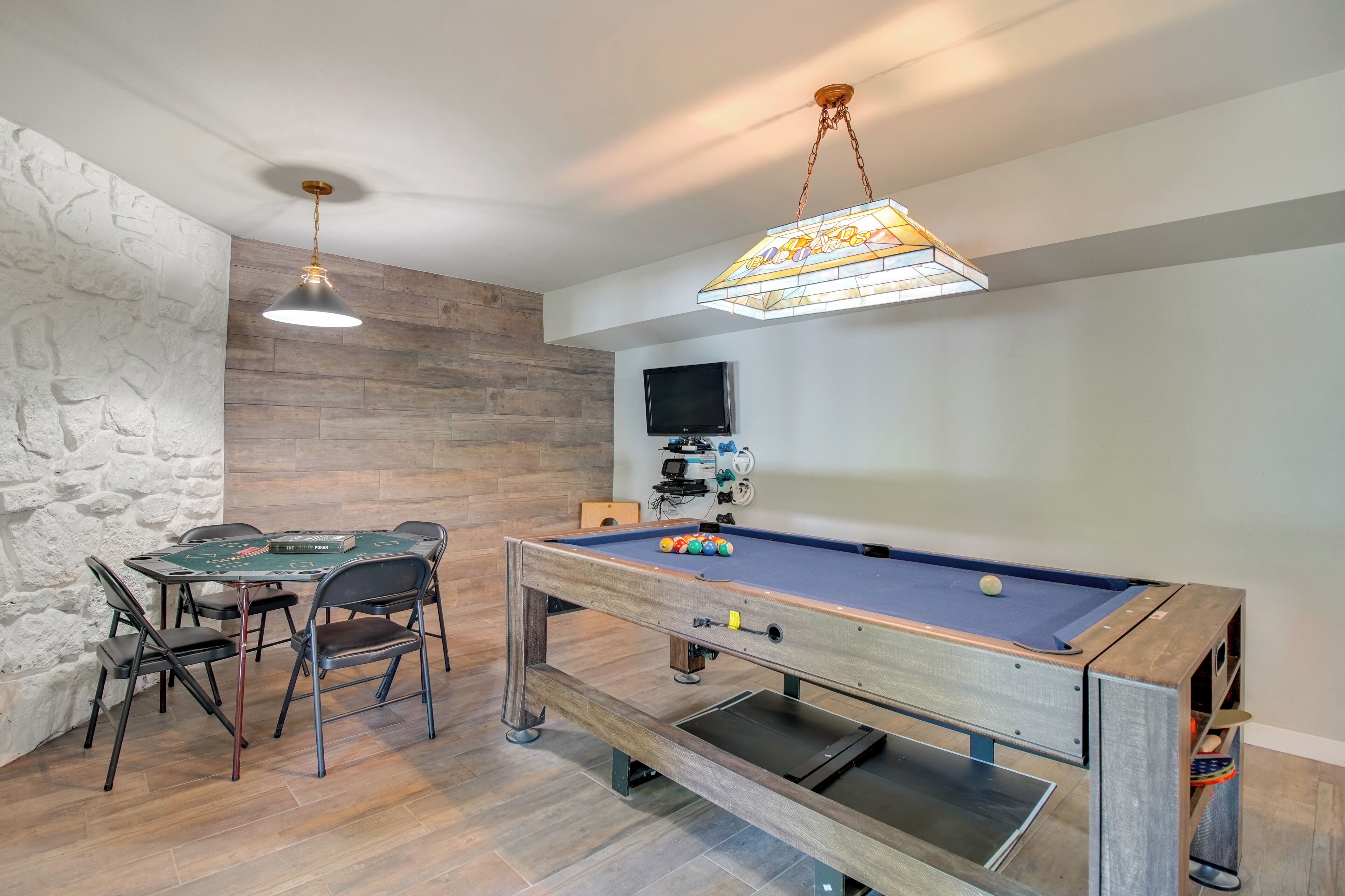 A pool table with chairs, a television mounted on the wall, and a stone-textured wall.