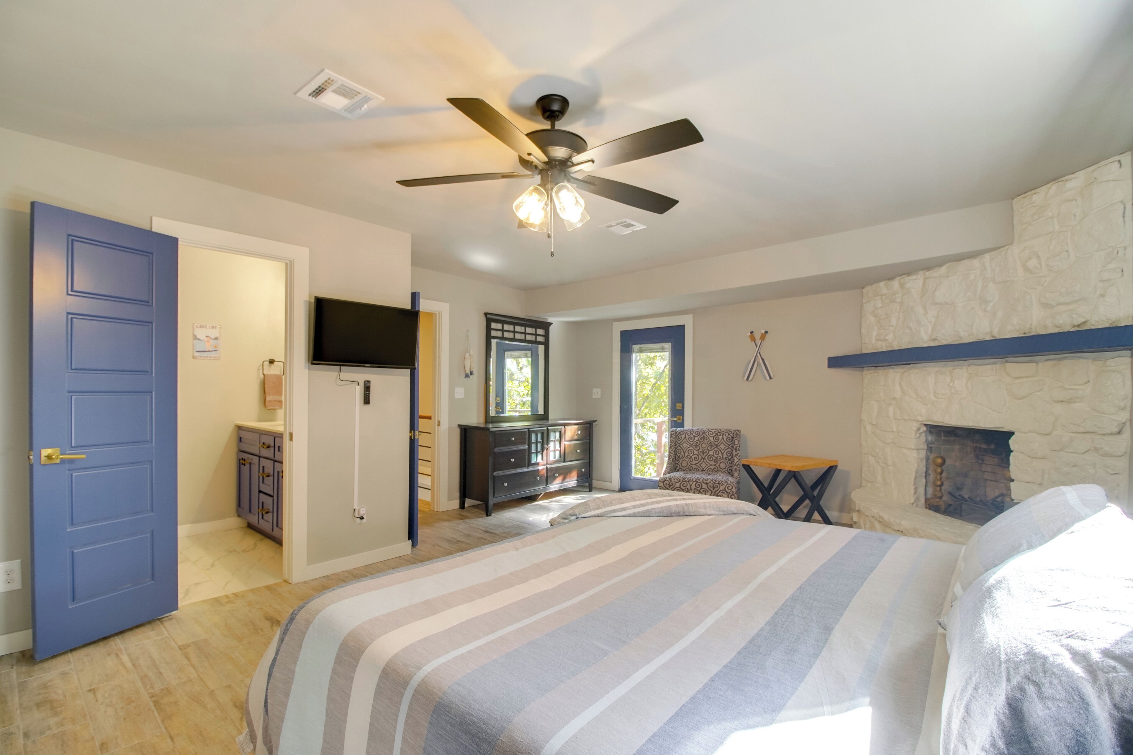 Une chambre à coucher comprenant un lit, un ventilateur de plafond, une cheminée en pierre, un téléviseur et une porte.