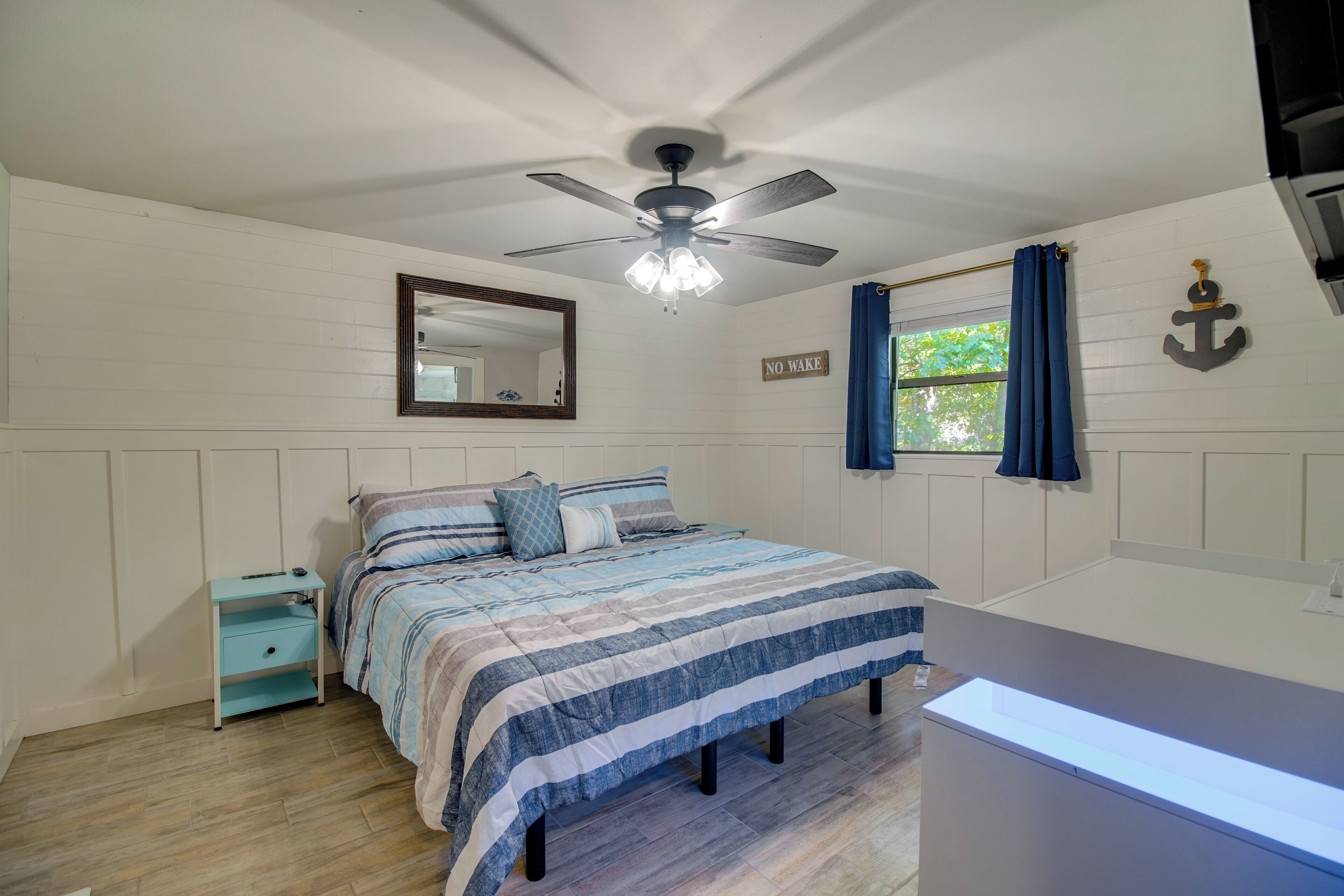 A bedroom with a bed, a nightstand, a ceiling fan, a window with curtains, a mirror, and a wall-mounted anchor decoration.