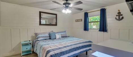 A bedroom with a bed, a nightstand, a ceiling fan, a window with curtains, a mirror, and a wall-mounted anchor decoration.