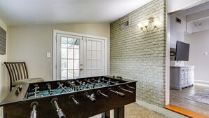 A room with a foosball table, a door, a brick wall, and a television in the background.