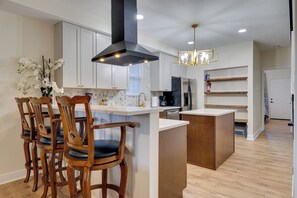A modern kitchen with white cabinets, a central island, wooden chairs, and a black range hood.