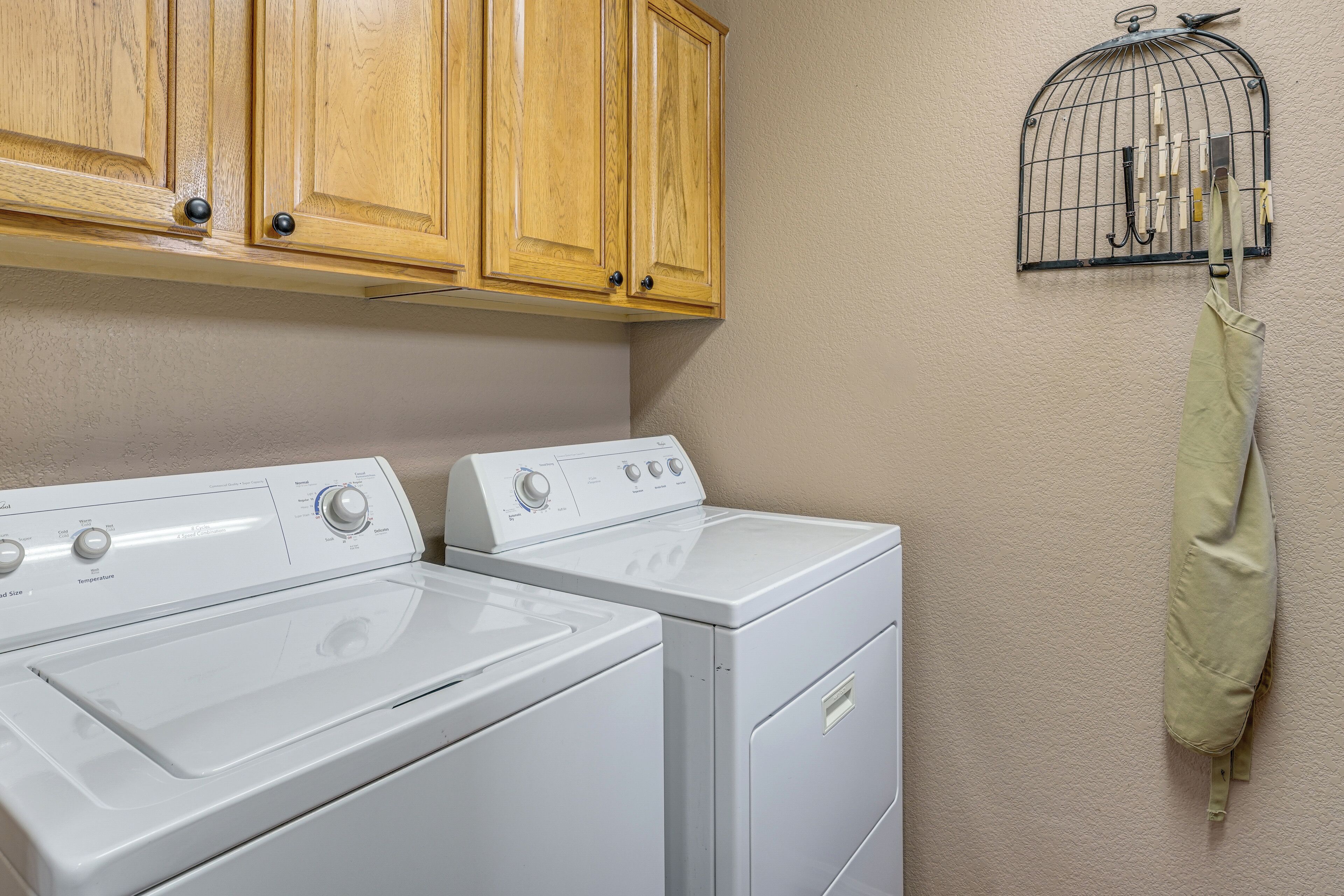 A laundry area with two white washing machines, wooden cabinets above, and a birdcage with a towel hanging on it.